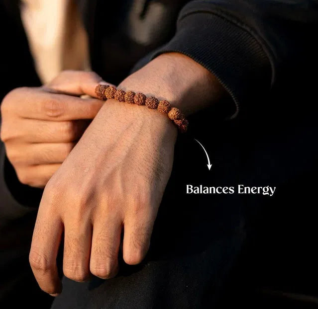 Hand wearing a rudraksha bracelet with text 'Balances Energy' on a dark background