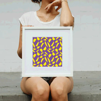 Person holding a framed abstract art print with purple and yellow pattern against a light gray background